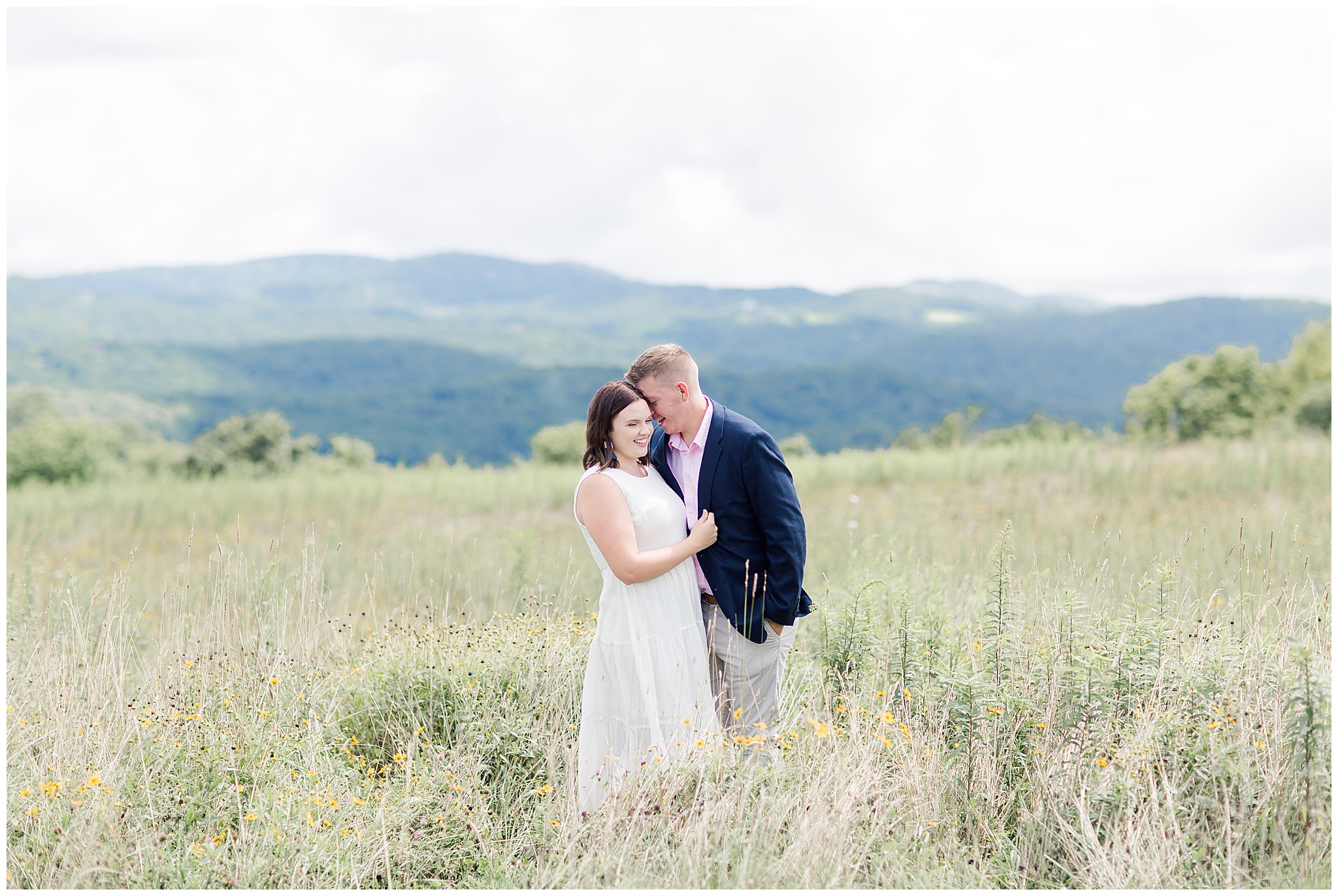 engaged couple in a field with the mountain scape behind them wind blowing in hair at moses cone park with girl in white dress navy suit jacket